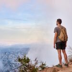 Full length shot of a young male hiker standing on a mountain trail looking through the clouds at the city below